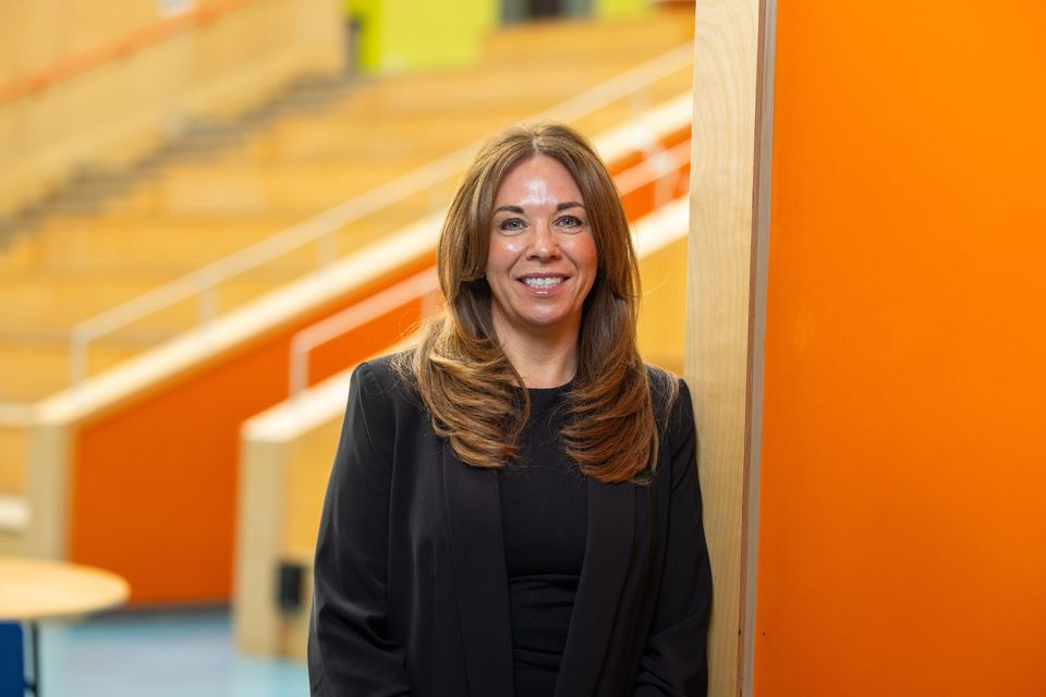 A person named Claire Madeloso with long hair is smiling and wearing a black outfit, standing in front of an orange and beige background.
