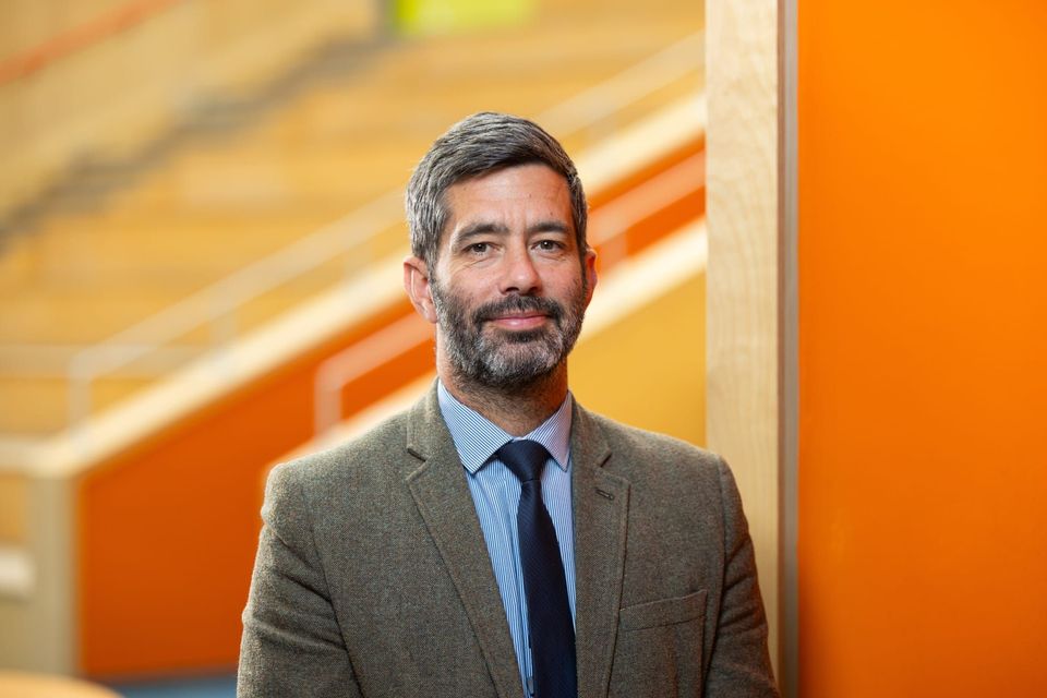 A man named Gary Joplin  with a gray beard, wearing a brown suit and blue tie, stands in front of an orange background with stairs in the distance.