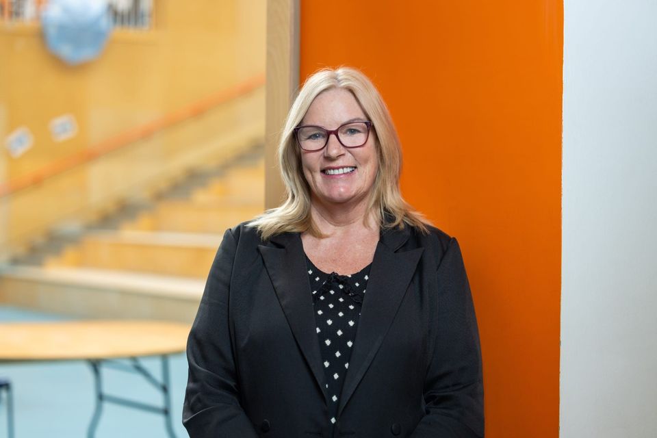 A woman named Paula Williams with blonde hair, wearing glasses and a black blazer, is standing and smiling in front of an orange wall and a staircase.
