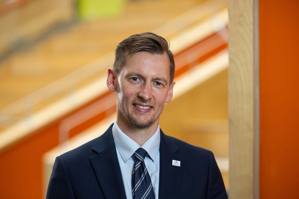 A man named Mark Ward in a suit is standing indoors, smiling at the camera with a blurred background.