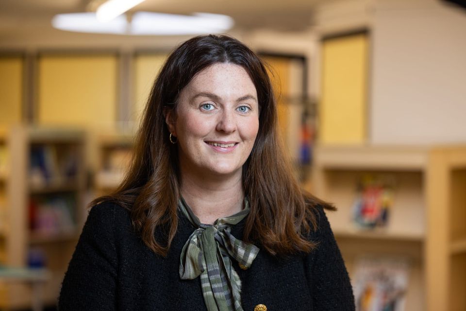 The image shows a person named Lyndsey Lewis smiling while standing in a room with shelves in the background. The image shows a person named Lyndsey Lewis smiling while standing in a room with shelves in the background.