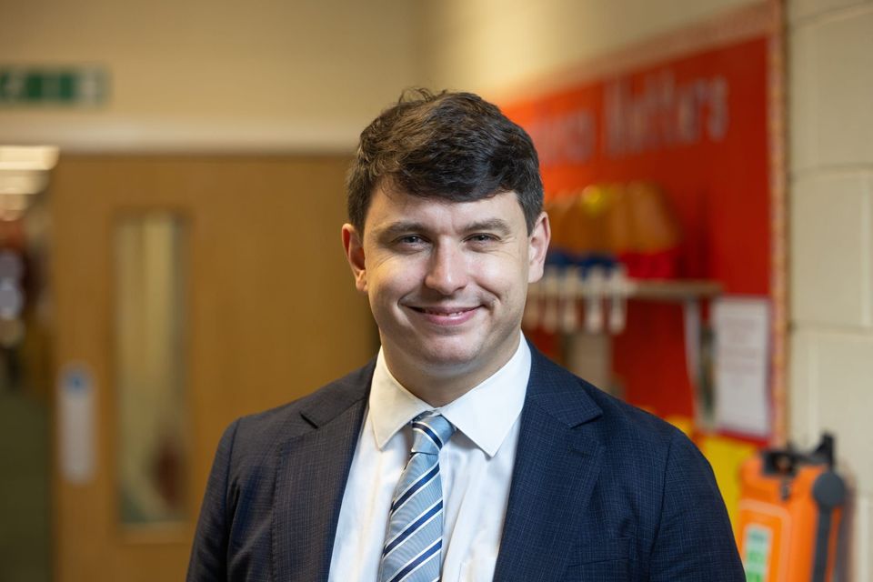 A man named Leon Tootle in a suit and tie is smiling in a hallway with a blurred background. A man named Leon Tootle in a suit and tie is smiling in a hallway with a blurred background.