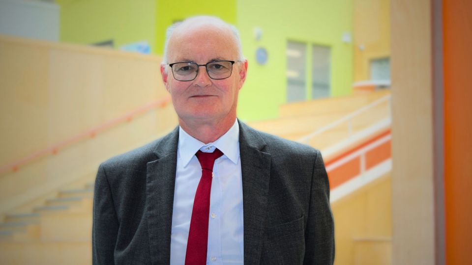A man named John Byrne in a suit and red tie is standing in a modern building with colorful walls.